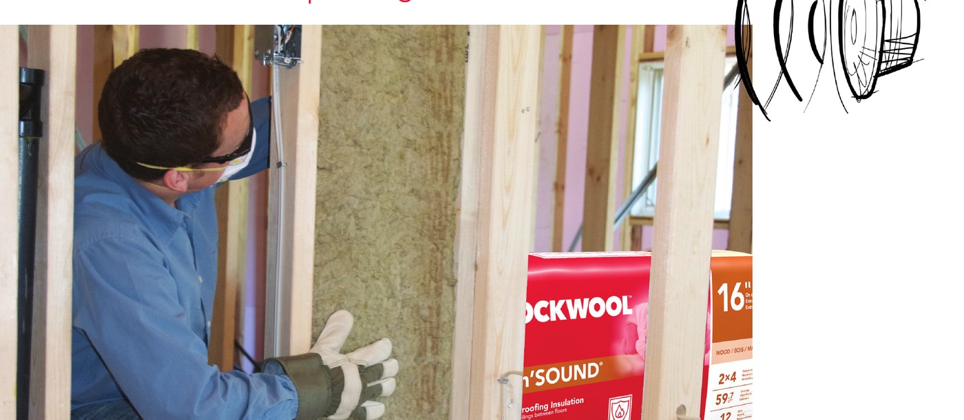 An installer pressing a stone wool batt into an interior wall with bags of Safe'n'Sound stacked beside the framing