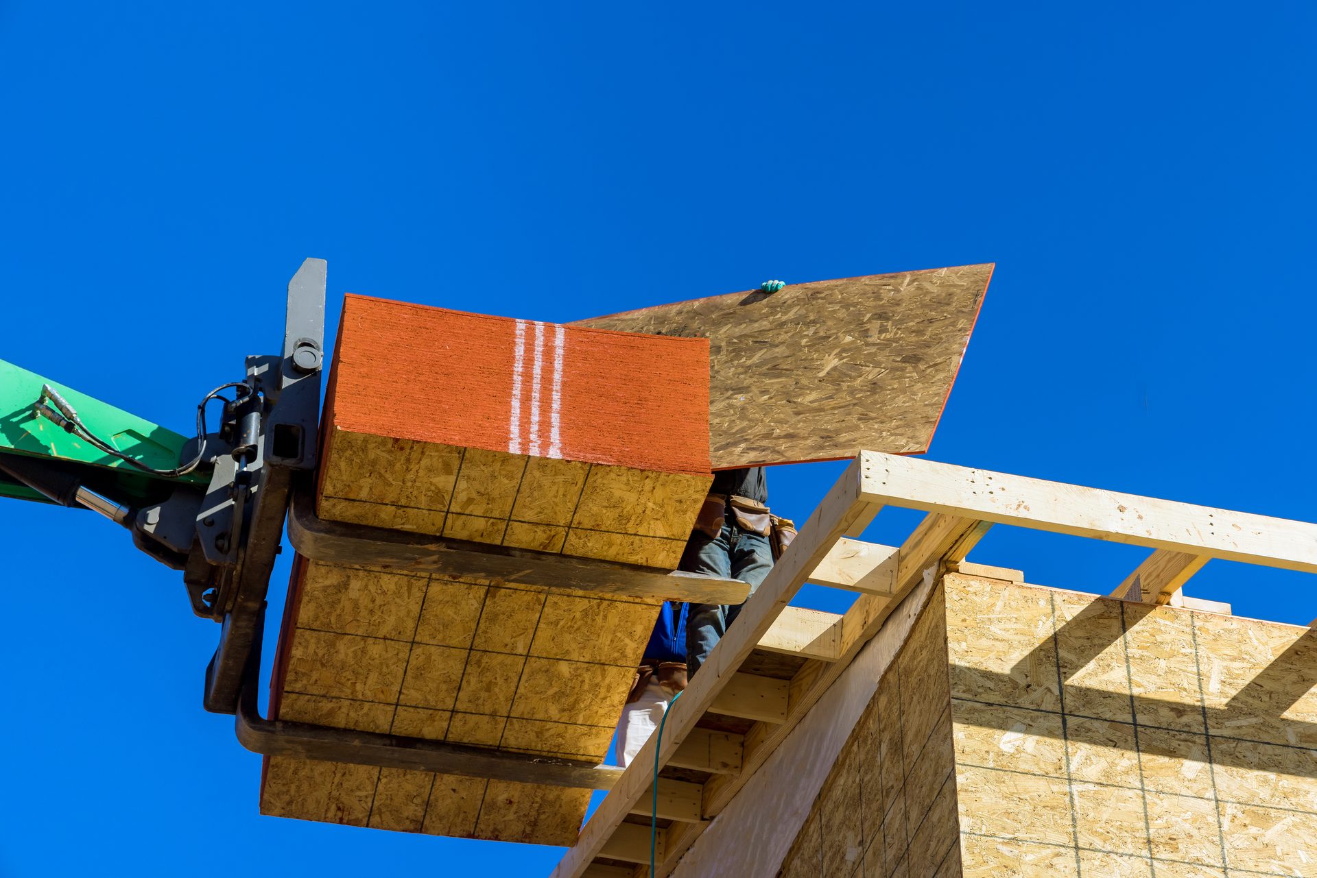 A forklift hoisting plywood and OSB sheathing onto a house under construction